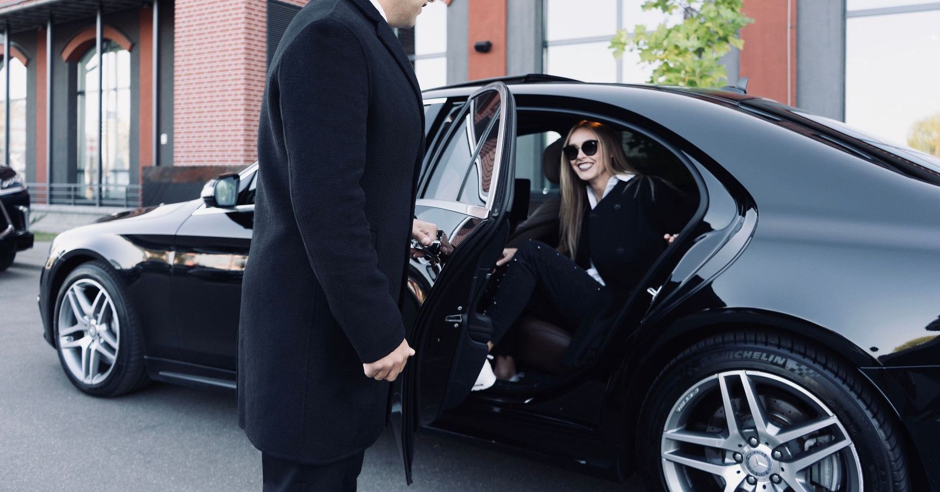 Man in coat smiling as he opens a black car door for a woman wearing sunglasses.